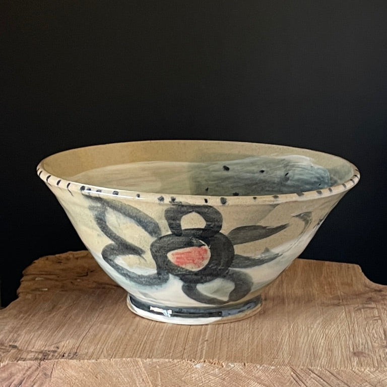 A handmade ceramic bowl with a hand-painted floral design in black, blue, and pink on a cream-colored glaze, displayed on a rustic wooden surface against a dark background.