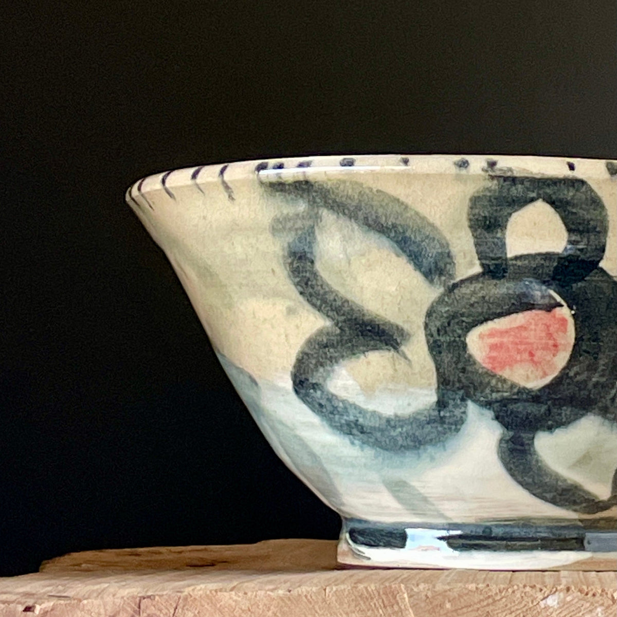 A handmade ceramic bowl with an abstract hand-painted floral pattern in black, blue, and pink on a cream-colored glaze, resting on a rustic wooden block against a dark background.