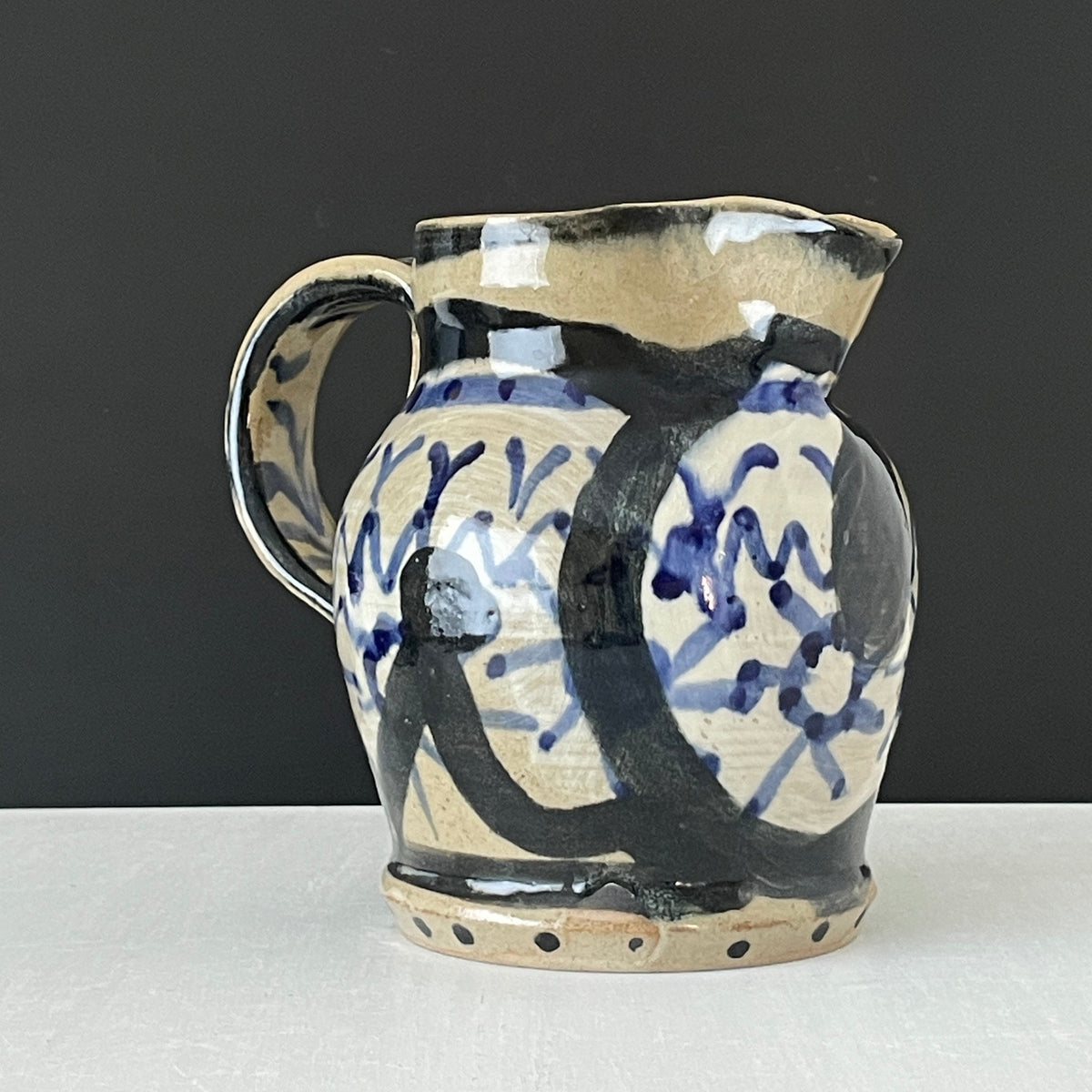 A handmade ceramic pitcher with a cream-colored glaze, decorated with bold black and blue abstract brushstrokes, set against a dark gray background and a light gray surface.